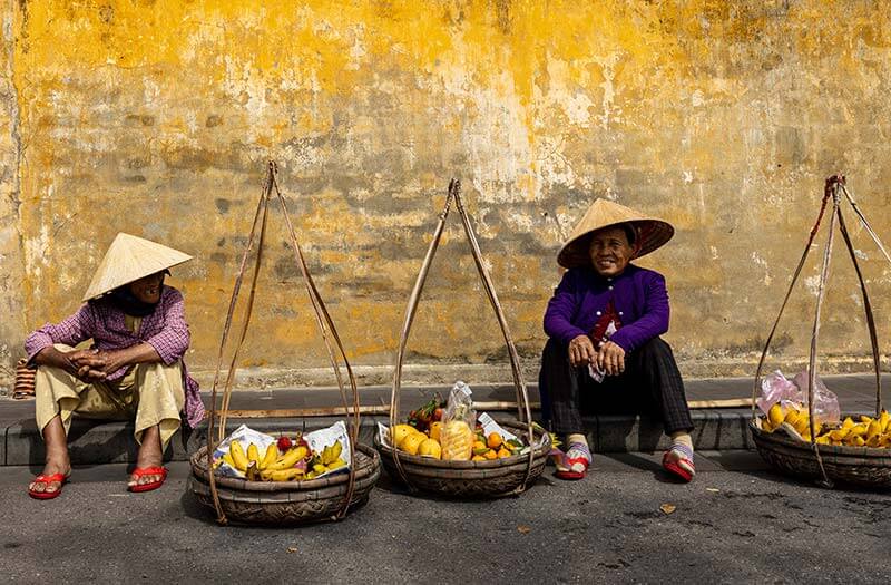 Two local vietnamese fruit sellers sitting on the side of a road with baskets of fruit.