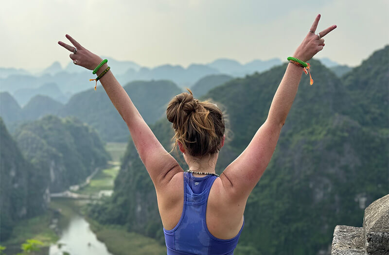 A woman on tour in Vietnam standing on a mountain top with her arms raised.